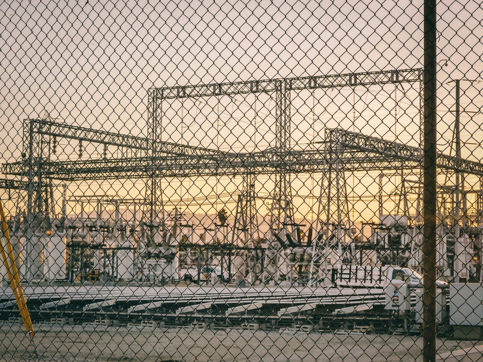 Photo by Kindel Media View of an industrial power plant through a chain-link fence at sunrise, symbolizing energy and industry.